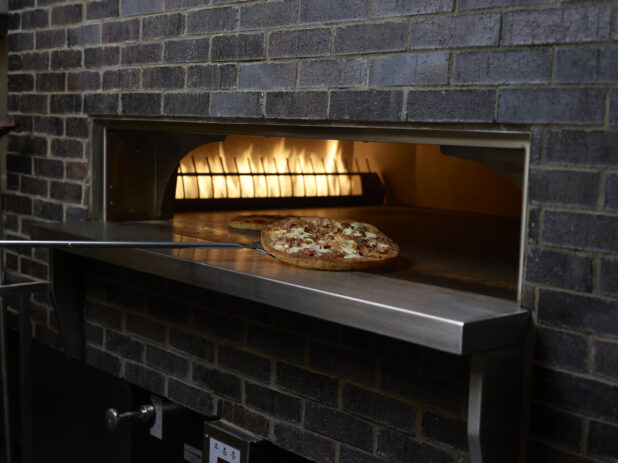 Whole Pizza on a Pizza Peel Coming Out of a Wood Fired Pizza Oven in a Restaurant Kitchen Setting