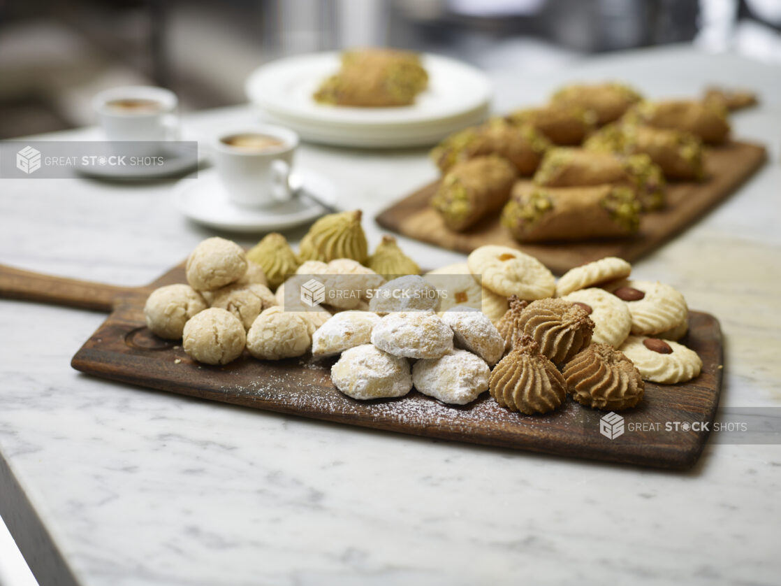 Assorted Italian Butter Cookies on a Wooden Platter Board with Pistachio Cannoli in the Background with Espresso Coffee in Ceramic Cups on a Marble Surface