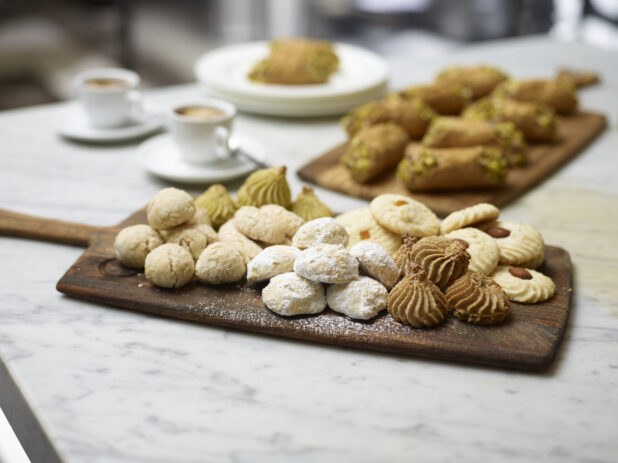 Assorted Italian Butter Cookies on a Wooden Platter Board with Pistachio Cannoli in the Background with Espresso Coffee in Ceramic Cups on a Marble Surface