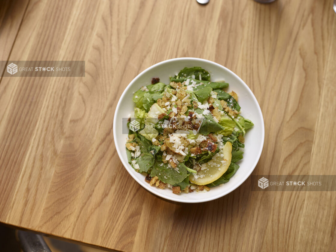 Classic Caesar Salad with a Lemon Wedge in a Round White Ceramic Salad Bowl on a Wooden Table