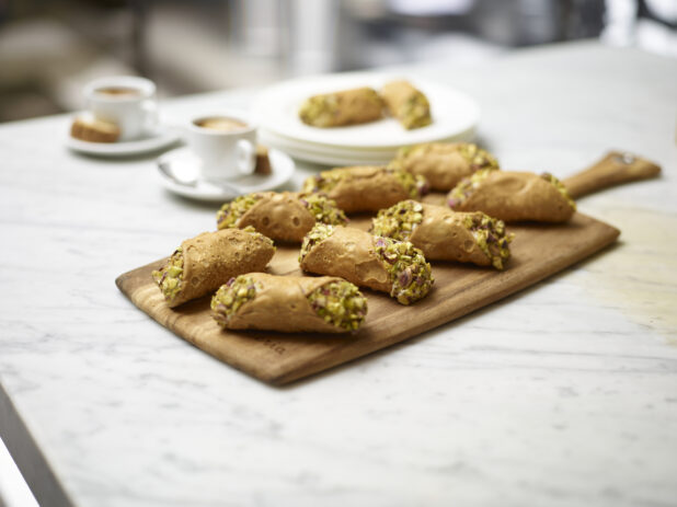 Close Up of Pistachio Cannolis on a Wooden Serving Boards with Espresso Coffee in Ceramic Cups on a White Marble Counter Top