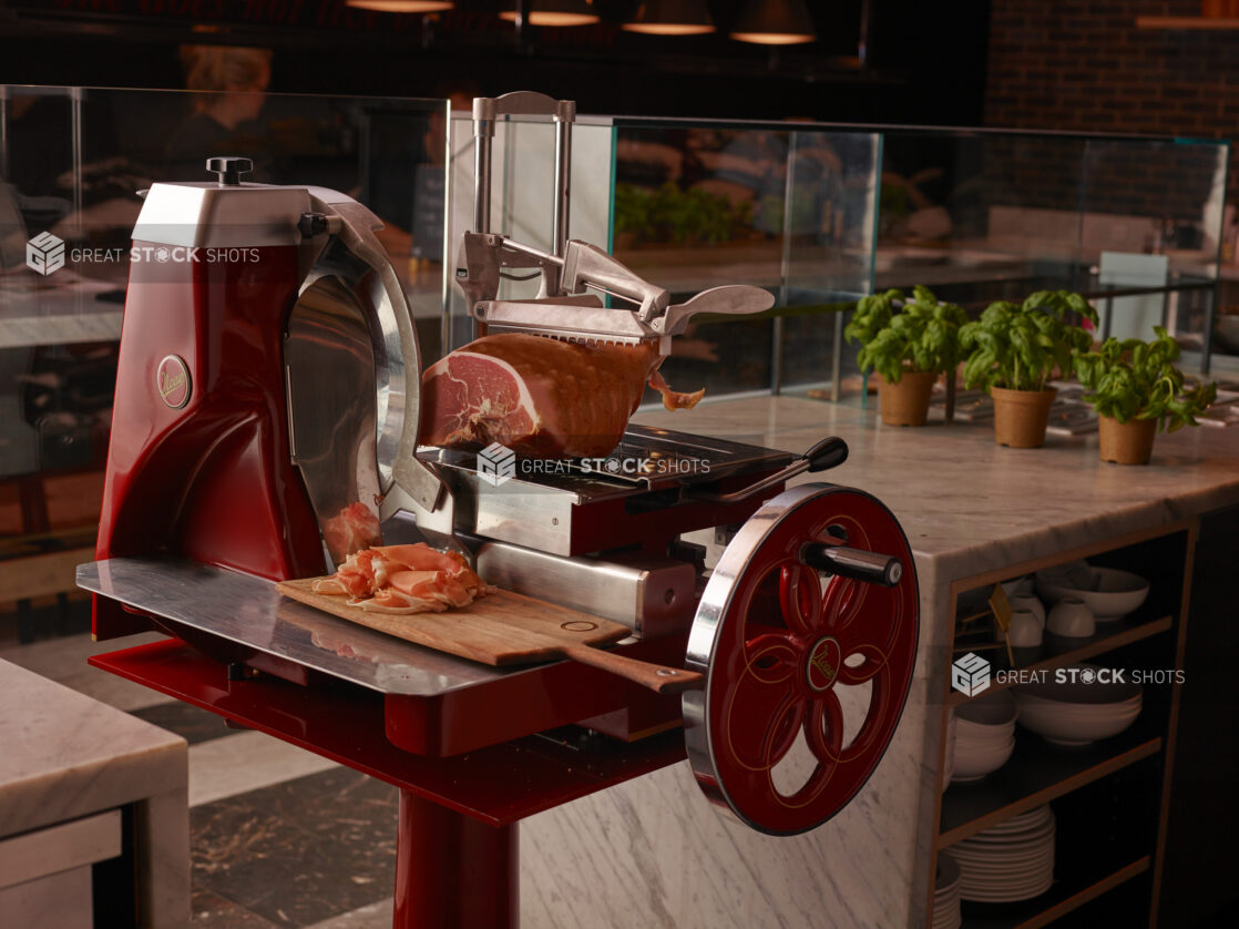 Fire Engine Red Berkel Flywheel Prosciutto Deli Meat Slicer with Slices of Prosciutto on a Wooden Serving Board in a Restaurant Interior Setting with Pots of Fresh Basil Leaves on Marble Counter Tops