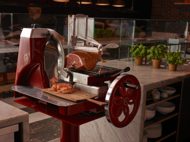 Fire Engine Red Berkel Flywheel Prosciutto Deli Meat Slicer with Slices of Prosciutto on a Wooden Serving Board in a Restaurant Interior Setting with Pots of Fresh Basil Leaves on Marble Counter Tops