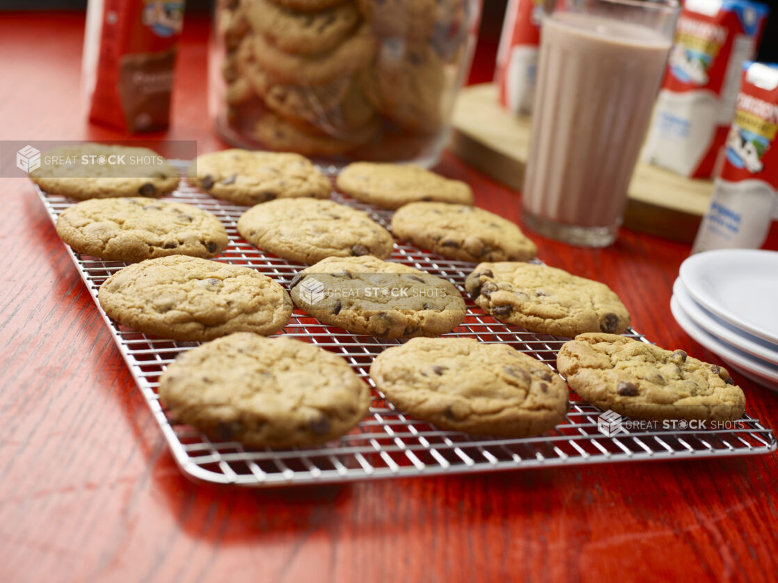 Tray of chocolate chip cookies cooling on a metal rack with a jar of cookies and chocolate milk in the background