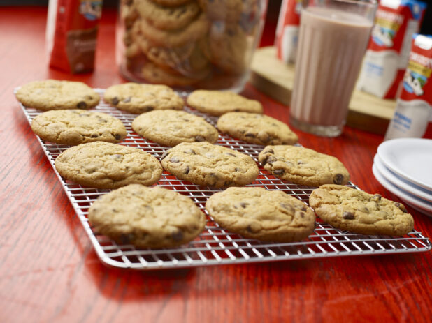 Tray of chocolate chip cookies cooling on a metal rack with a jar of cookies and chocolate milk in the background