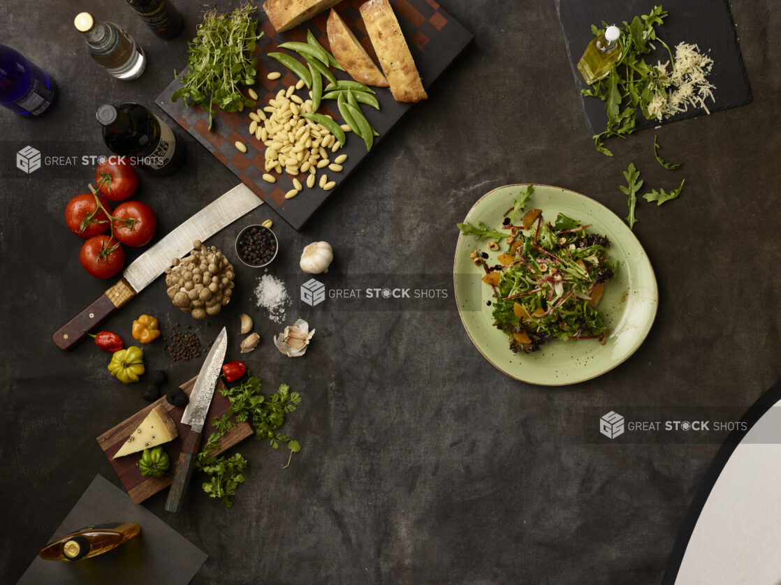 Bowl of arugula salad and many fresh ingredients on a dark grey cloth background