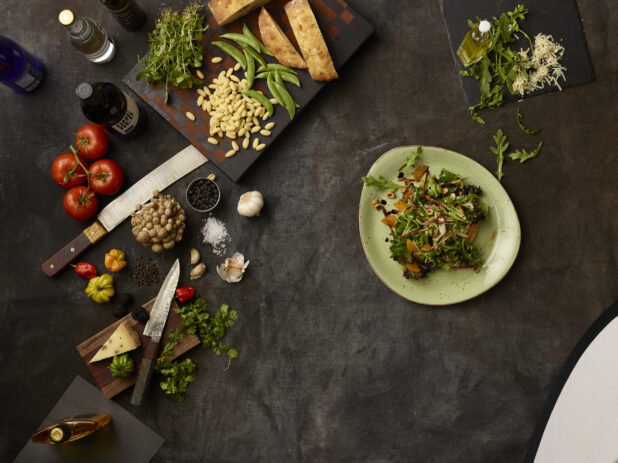 Bowl of arugula salad and many fresh ingredients on a dark grey cloth background