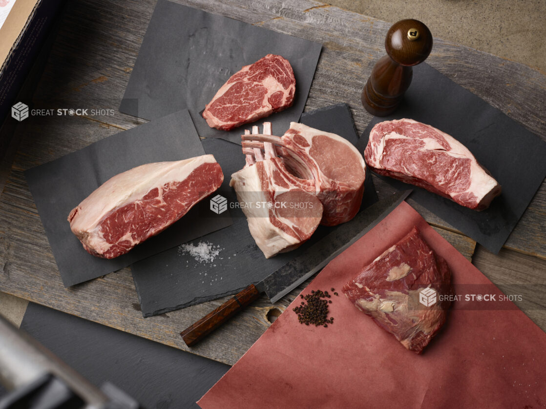 Assorted cuts of meat, steak, chops with salt and peppercorns on butcher paper on a wood background