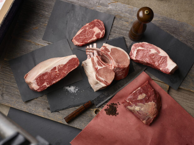 Assorted cuts of meat, steak, chops with salt and peppercorns on butcher paper on a wood background