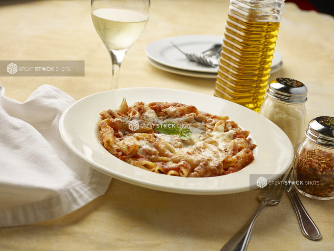 Bowl of penne pasta in tomato sauce with melted cheese on top with a glass of white wine on a yellow background