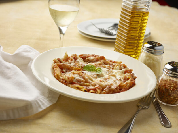 Bowl of penne pasta in tomato sauce with melted cheese on top with a glass of white wine on a yellow background