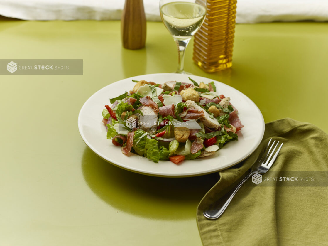 Chopped salad with prosciutto, chicken, salami, red onion, red peppers and shaved parmesan with a glass of white wine on a green background