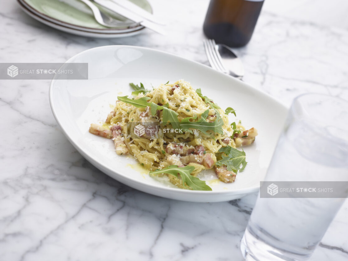 Bowl of spaghetti carbonara garnished with arugula and grated parmesan cheese on a white marble table