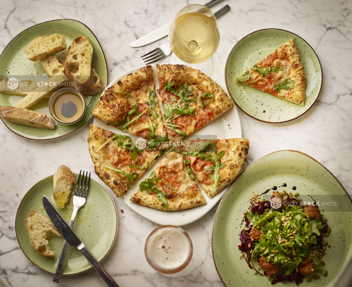 Overhead view of sliced wood fire pepperoni pizza, bread and an arugula salad with chicken on green plates on a white marble background