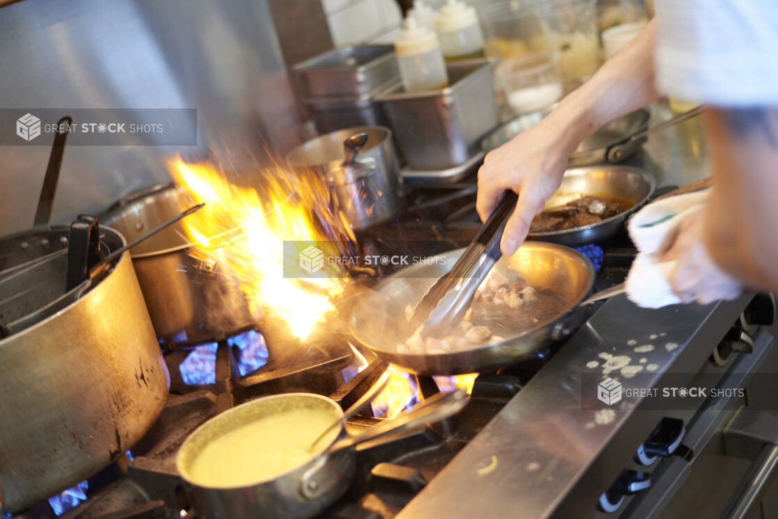 Chef's hands with tongs and a sauté pan over a busy gas range with orange and blue flames