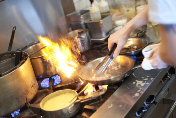 Chef's hands with tongs and a sauté pan over a busy gas range with orange and blue flames