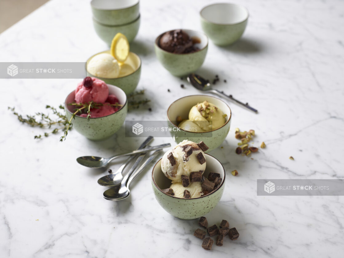 A variety of sherbet, sorbet, and ice cream in small green speckled bowls with spoons and fresh ingredients on a white marble countertop