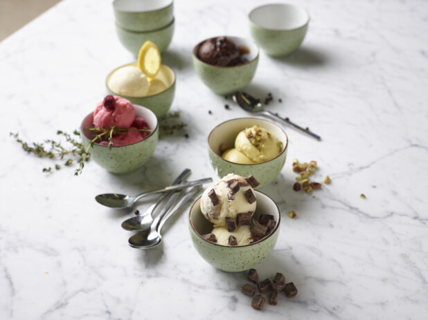 A variety of sherbet, sorbet, and ice cream in small green speckled bowls with spoons and fresh ingredients on a white marble countertop