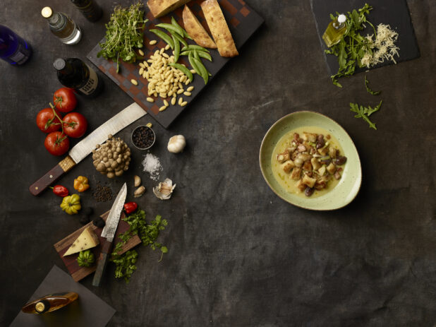 Bowl of gnocchi and many fresh ingredients on a dark grey cloth background