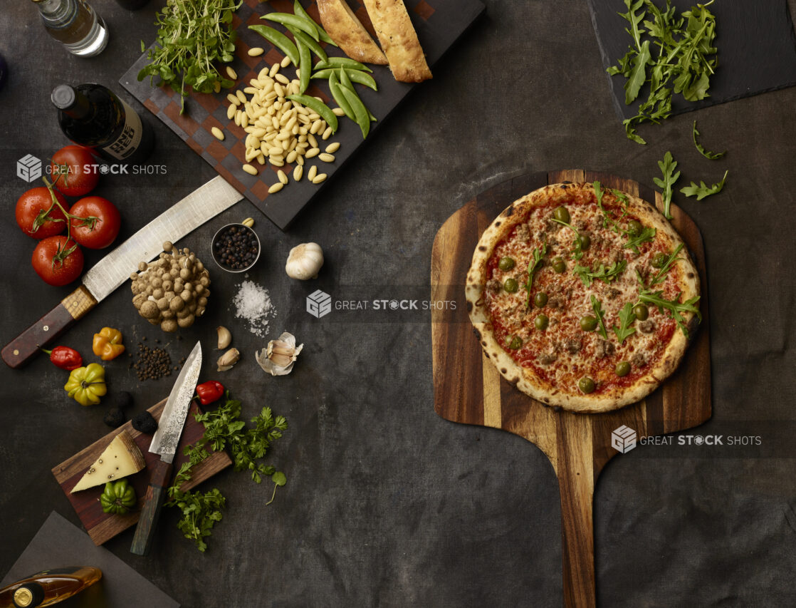 Pizza with olives and sausage surrounded by many fresh ingredients on a dark grey cloth background