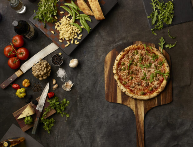 Pizza with olives and sausage surrounded by many fresh ingredients on a dark grey cloth background
