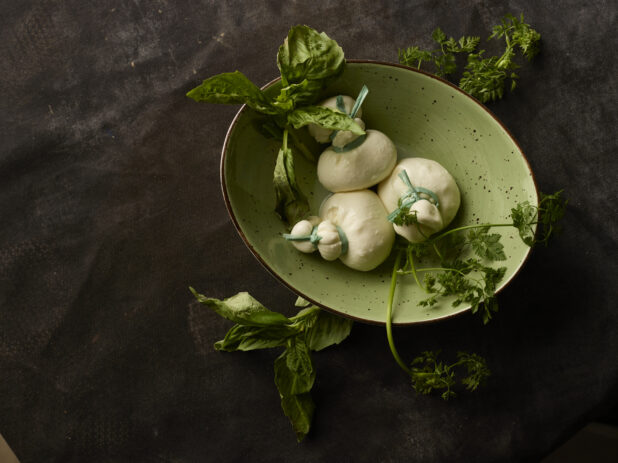 Three balls of Fresh Mozzarella Treccione in a green bowl surrounded by fresh herbs