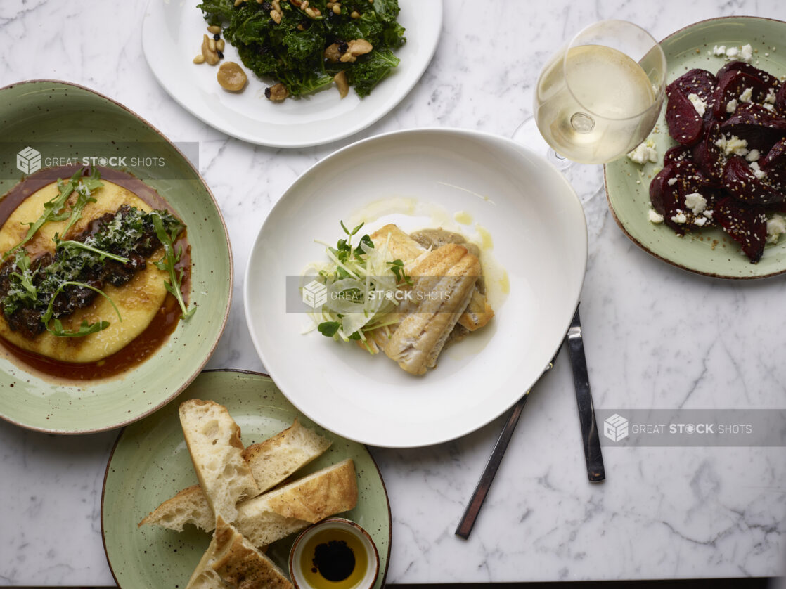 Two fillets of white fish, drizzled with butter and garnished with microgreens, other dishes surrounding, white marble background