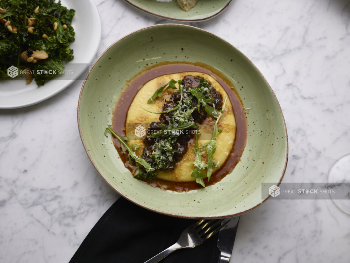 Overhead View of Braised Beef Short Ribs and Baby Arugula on a Bed of Polenta with Au Jus in a Green Ceramic Dish with a Side of Sautéed Kale on a Marble Table