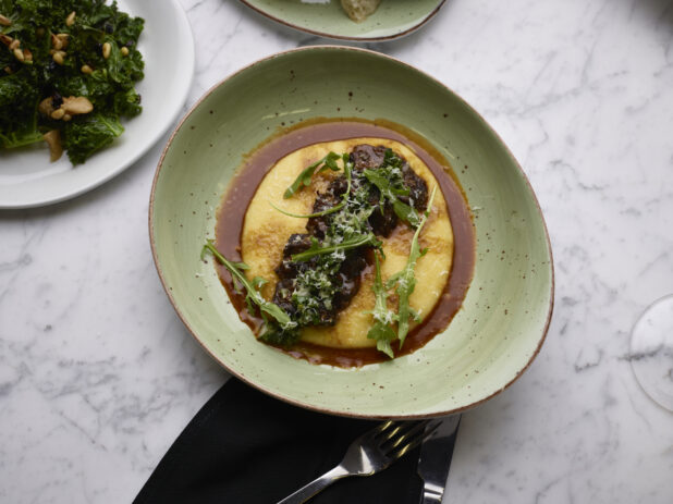 Overhead View of Braised Beef Short Ribs and Baby Arugula on a Bed of Polenta with Au Jus in a Green Ceramic Dish with a Side of Sautéed Kale on a Marble Table