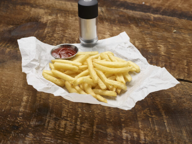 French fries with a ramekin of ketchup on parchment paper with a salt shaker on a rustic wooden background