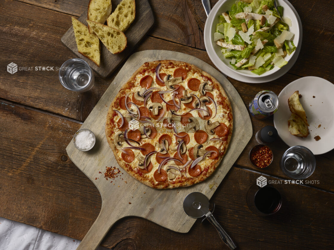 Whole 3 topping pizza with pepperoni, red onions and mushrooms on a wooden peel with a pizza cutter, caesar salad, garlic bread, soda can, on a rustic wooden background