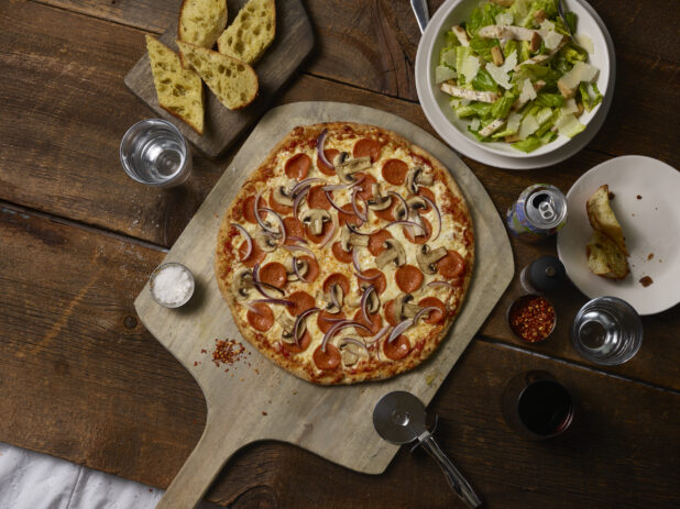 Whole 3 topping pizza with pepperoni, red onions and mushrooms on a wooden peel with a pizza cutter, caesar salad, garlic bread, soda can, on a rustic wooden background
