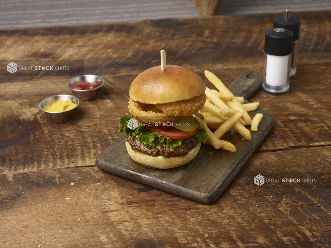 Hamburger with lettuce, tomato, pickles and onion rings with french fries on a wooden board with ramekins of ketchup and mustard and a salt shaker and pepper grinder in the background on a rustic wooden background