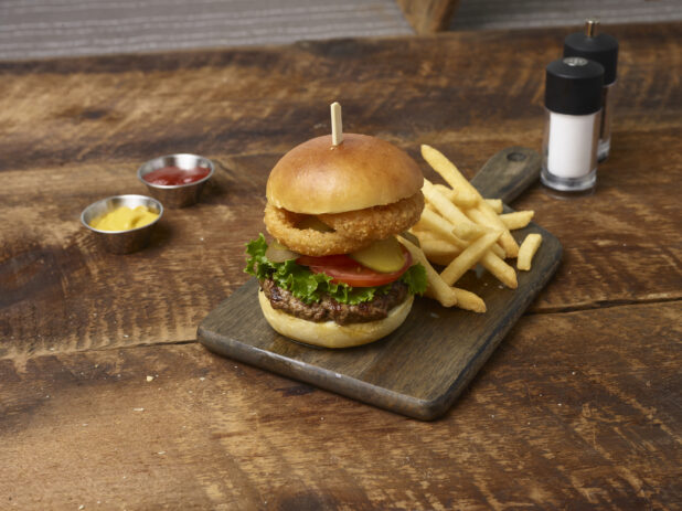 Hamburger with lettuce, tomato, pickles and onion rings with french fries on a wooden board with ramekins of ketchup and mustard and a salt shaker and pepper grinder in the background on a rustic wooden background