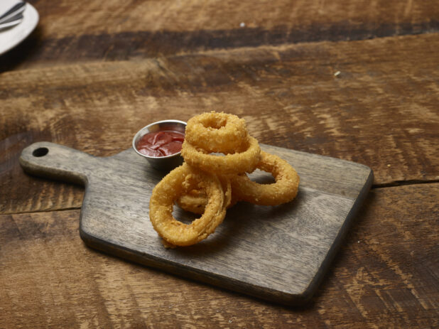 Fried onion rings with a ramekin of ketchup on a wooden board on a rustic wooden background
