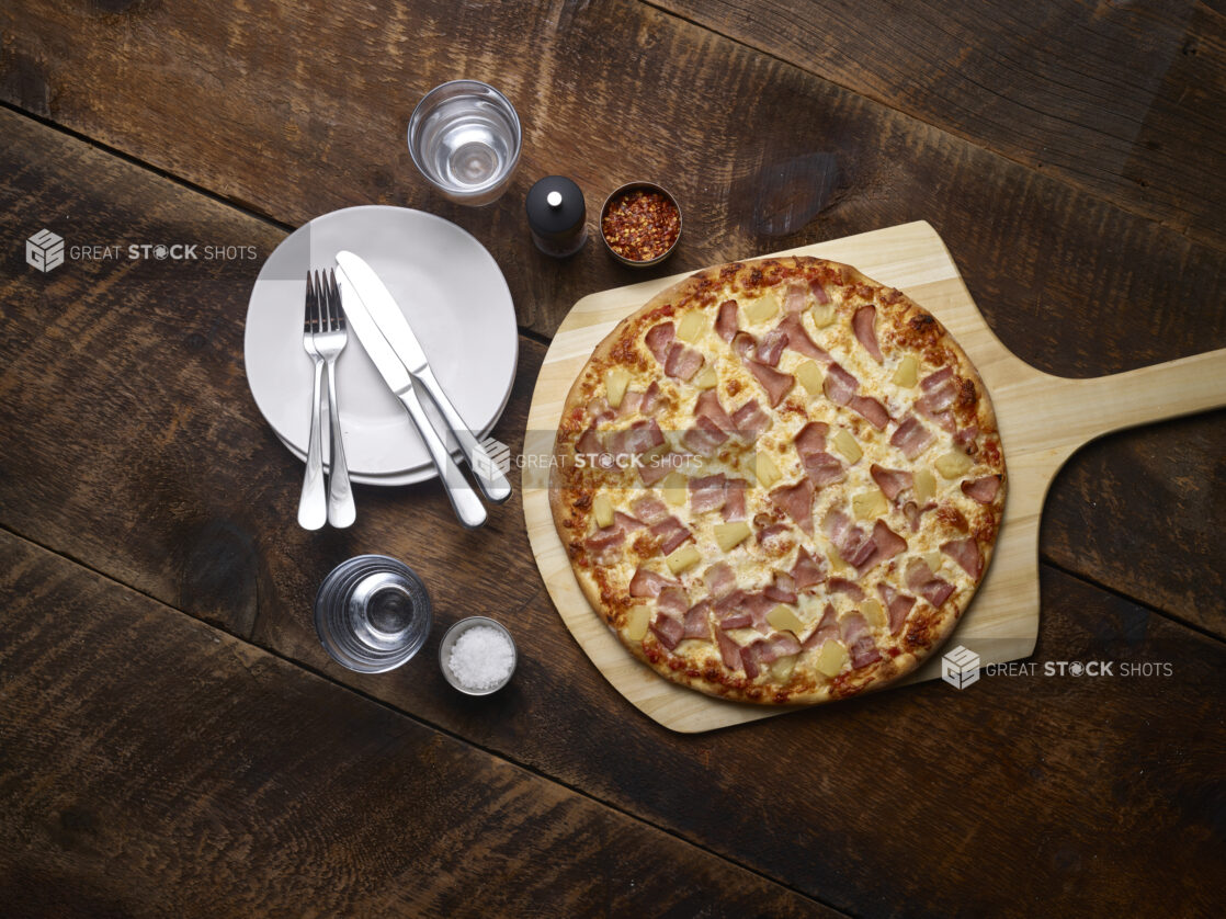 Whole Hawaiian pizza on a pizza peel with white side plates with cutlery, salt, chili flakes and a pepper mill on the side on a rustic wooden background