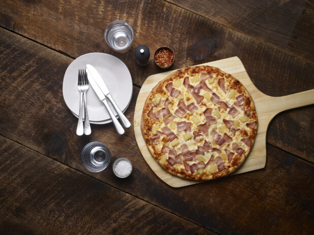 Whole Hawaiian pizza on a pizza peel with white side plates with cutlery, salt, chili flakes and a pepper mill on the side on a rustic wooden background