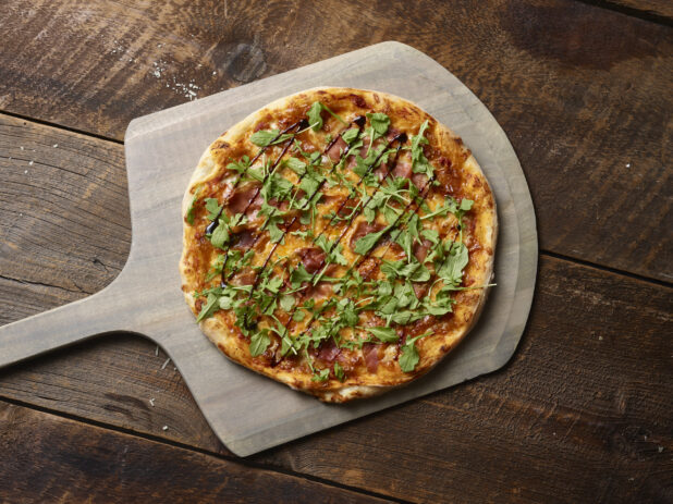 Whole thin crust pizza with arugula and prosciutto on a wood pizza peel, overhead shot