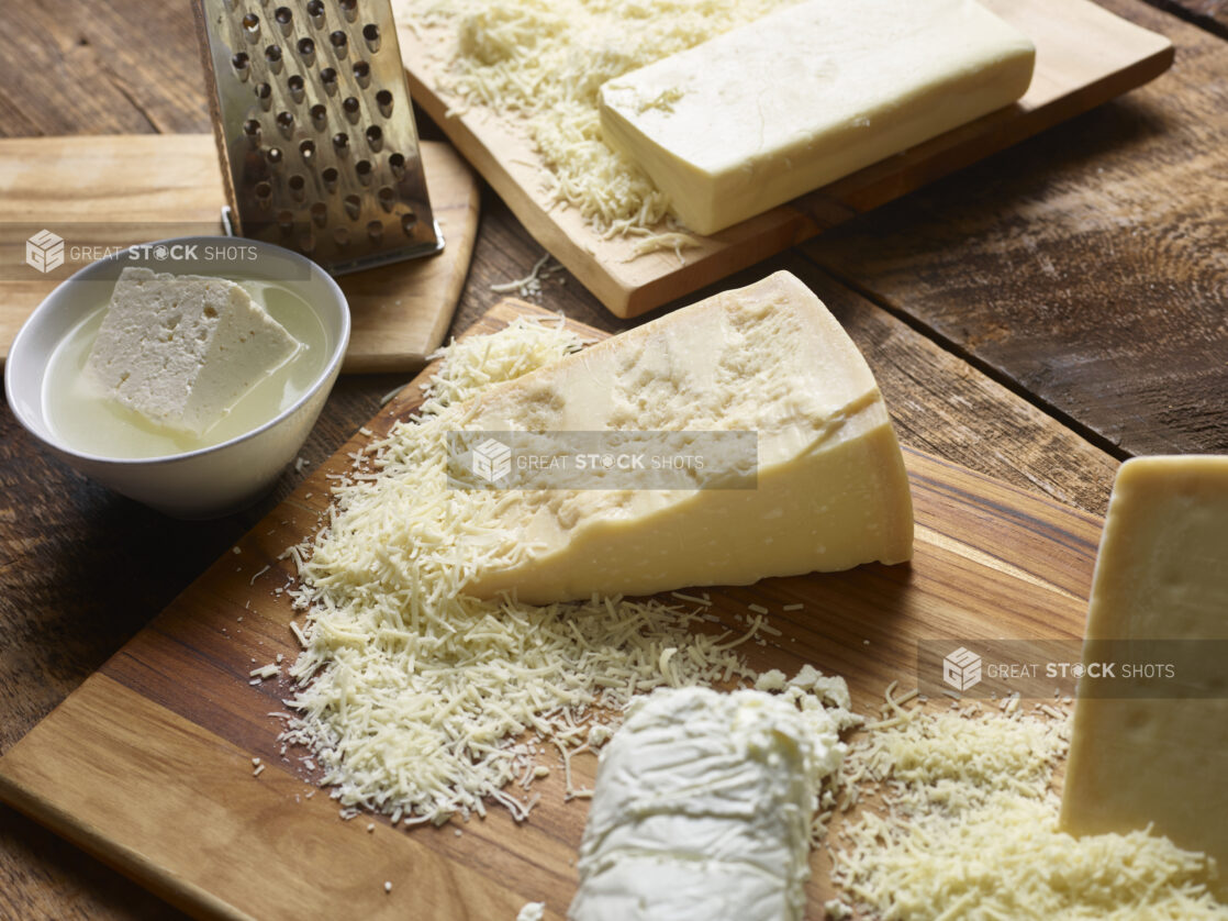 Blocks of partially grated white cheeses on cutting boards with a metal cheese grater, bowl with feta in brine