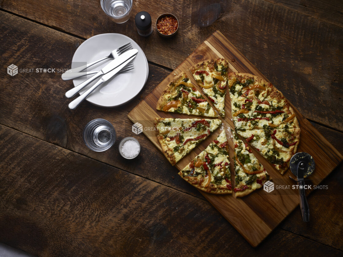 2 topping pizza sliced with roasted bell peppers and spinach on a wooden board with a pizza cutter with white side plates, cutlery, chili flakes, coarse salt and pepper mill