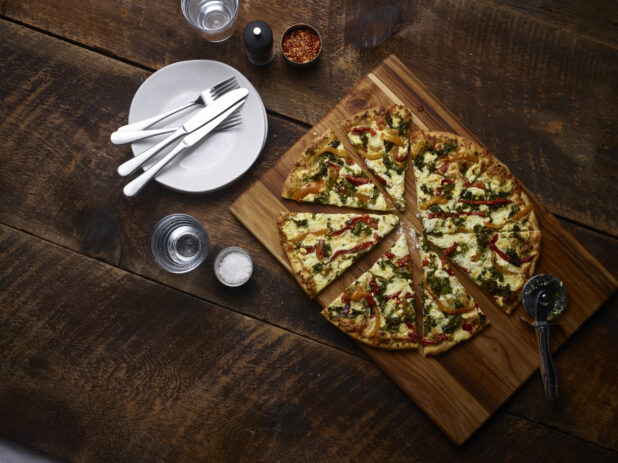 2 topping pizza sliced with roasted bell peppers and spinach on a wooden board with a pizza cutter with white side plates, cutlery, chili flakes, coarse salt and pepper mill