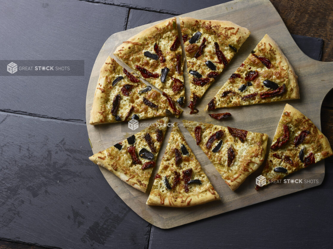 Whole 2 topping pizza with sundried tomatoes and black olives, sliced on a wooden peel on a slate background