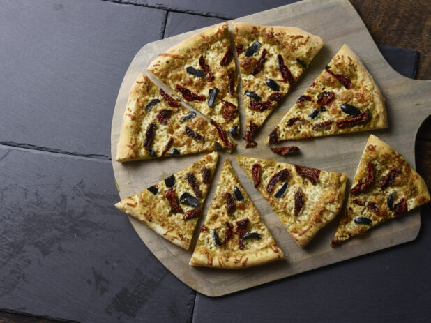 Whole 2 topping pizza with sundried tomatoes and black olives, sliced on a wooden peel on a slate background