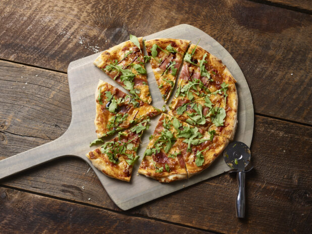 Whole thin crust pizza, sliced, with arugula and prosciutto on a wood pizza peel, overhead shot