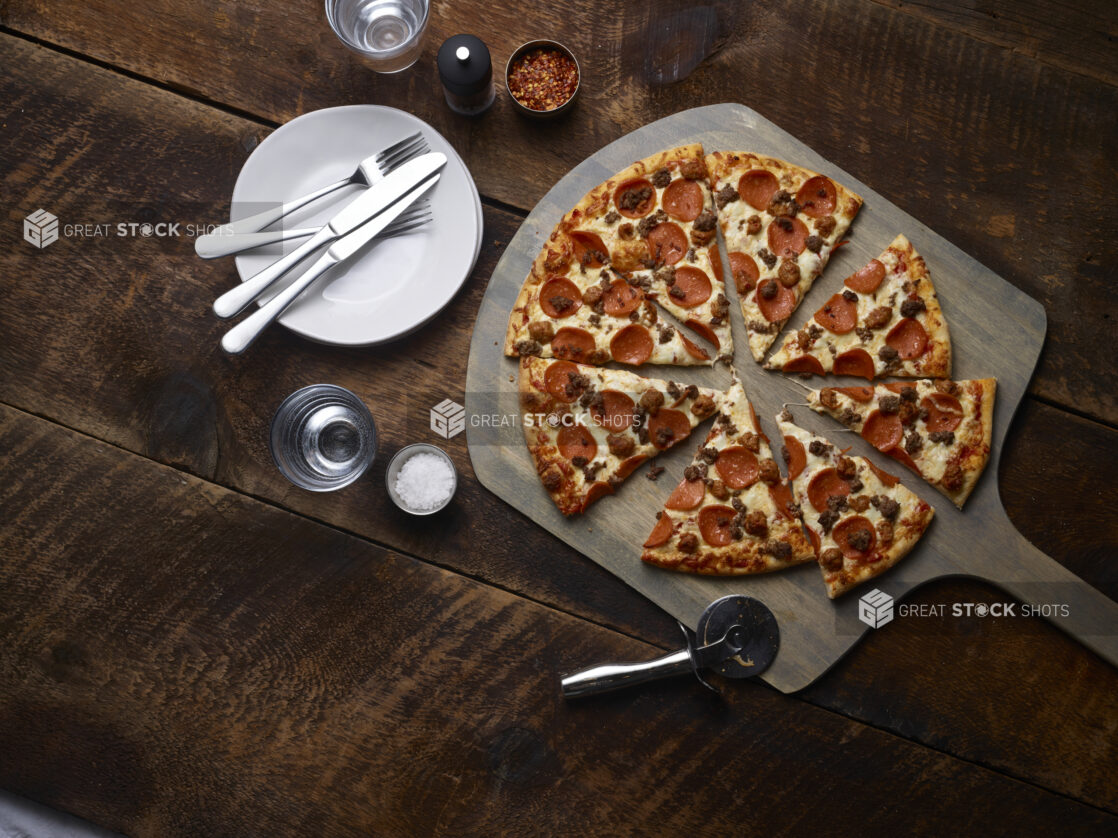 Whole 3 topping Meat Lovers pizza sliced with pepperoni, sausage and ground beef on a pizza peel with white side plates, cutlery, chili flakes, salt and pepper mill on a rustic wooden background