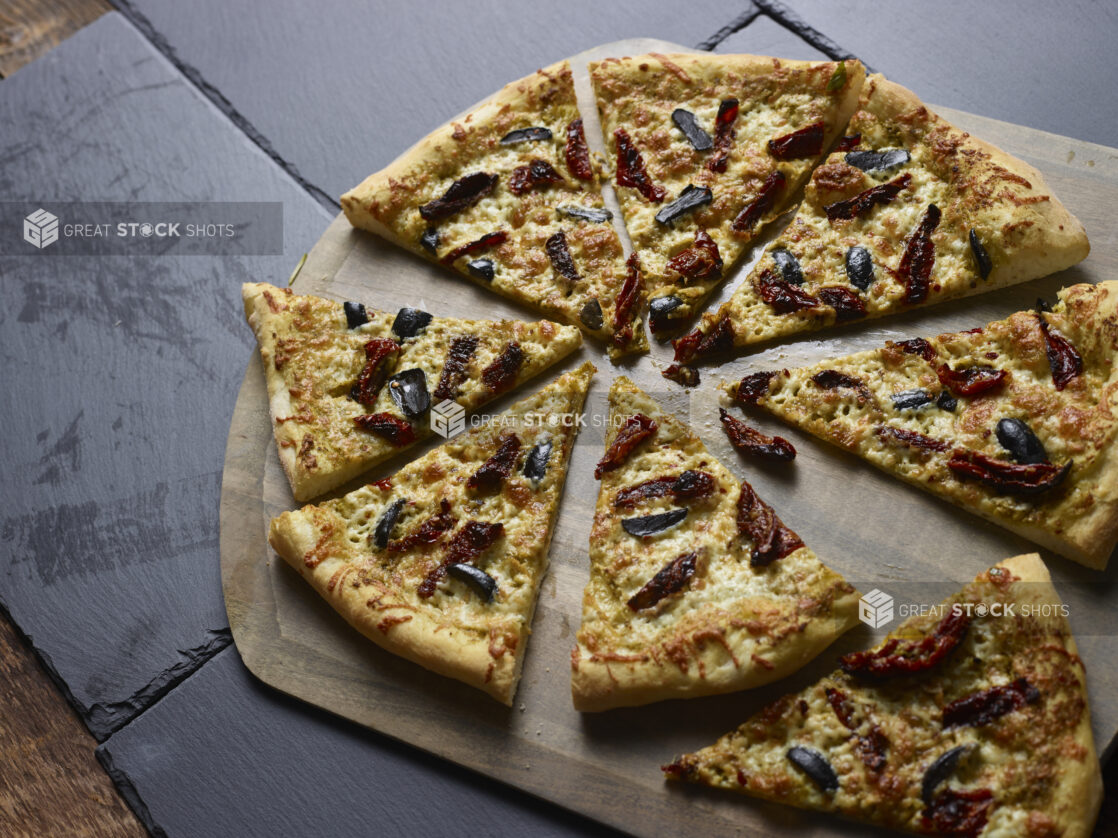 Whole 2 topping pizza with sundried tomatoes and black olives cut into slices on a wooden pizza peel, on a slate background