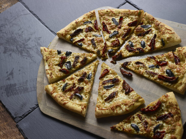 Whole 2 topping pizza with sundried tomatoes and black olives cut into slices on a wooden pizza peel, on a slate background