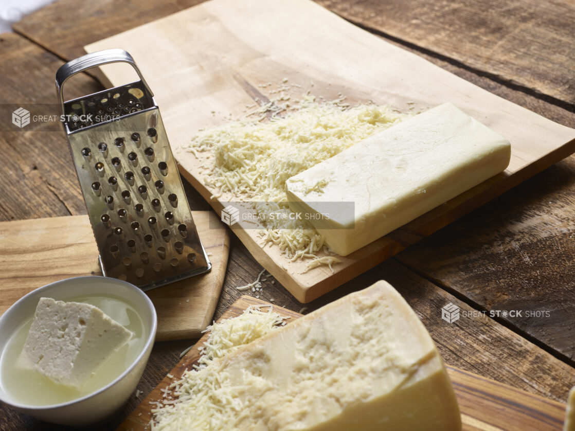 Blocks of white cheese on cutting boards with a steel cheese grater, bowl of feta cheese in brine