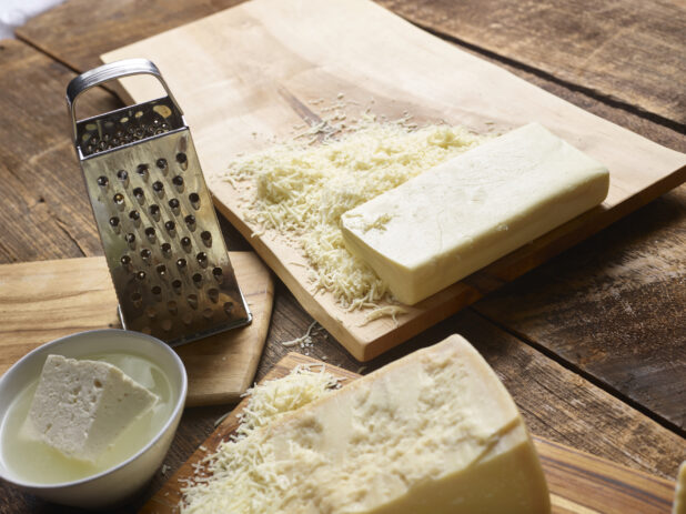 Blocks of white cheese on cutting boards with a steel cheese grater, bowl of feta cheese in brine