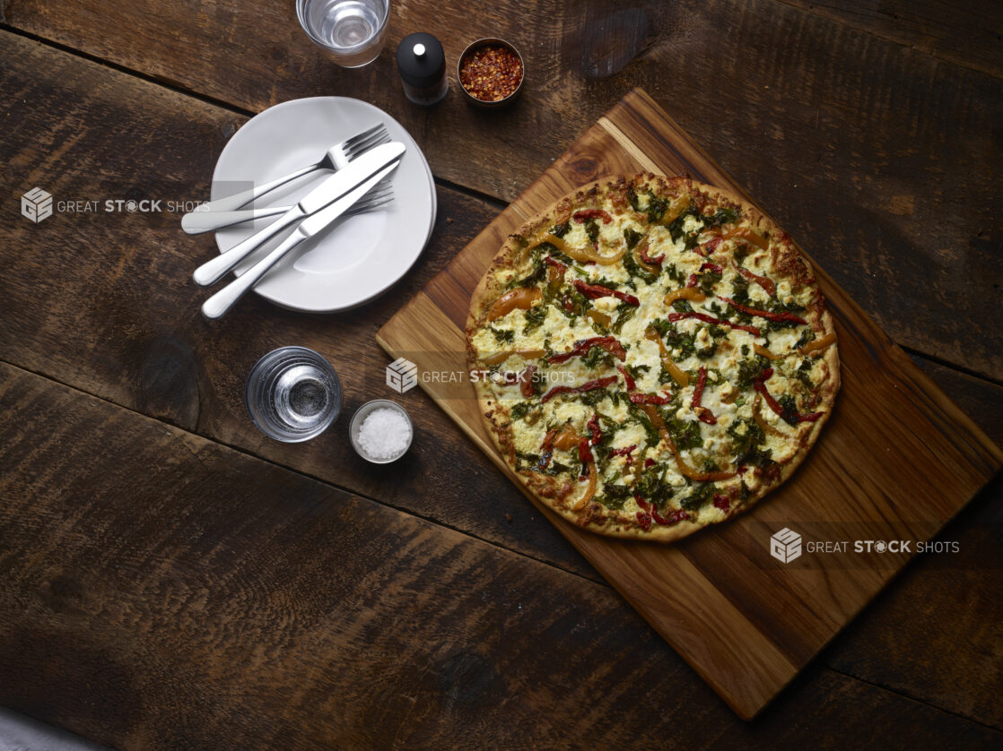 Whole 2 topping pizza with roasted bell peppers and spinach on a wooden board with white side plates, cutlery, chili flakes, coarse salt and pepper mill